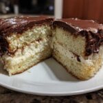 Cross-section view of a slice of Italian love cake showing two layers of light cake, creamy filling, and thick chocolate frosting.
