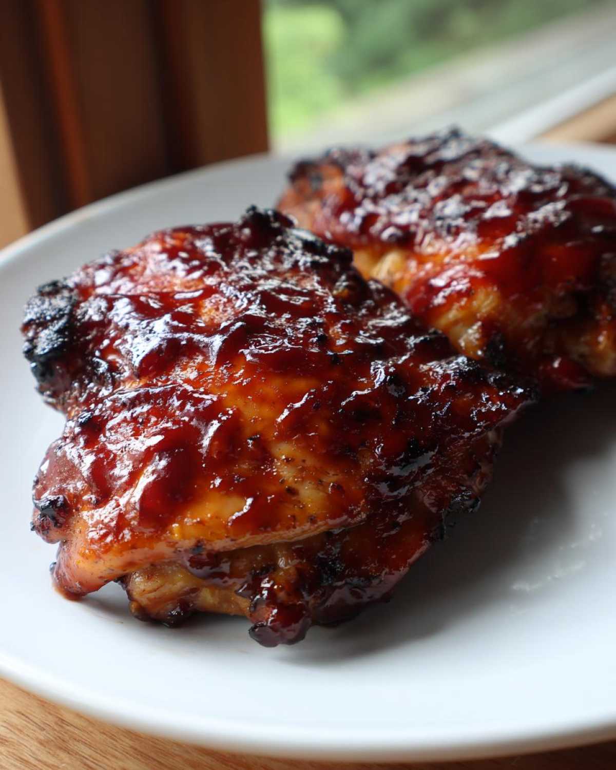 Two pieces of juicy bbq chicken thighs covered in a thick, glossy, dark red barbecue sauce, served on a white plate.