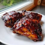 Three pieces of juicy, glazed bbq chicken thighs with a dark, caramelized crust served on a white plate.