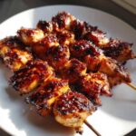 Close-up of several juicy chicken skewers with a dark, caramelized glaze, resting on a white plate.