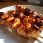 Close-up of three skewers featuring juicy, glazed, and slightly charred chicken skewers resting on a white plate.
