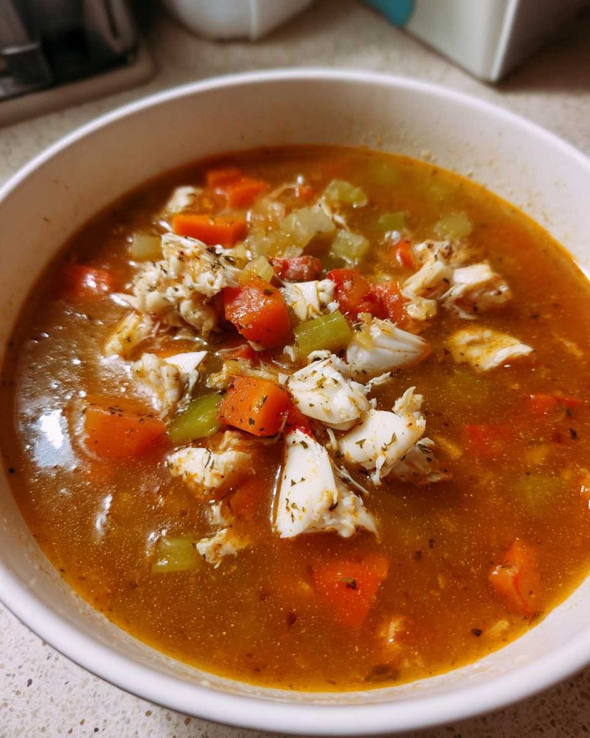 Close-up of a hearty bowl of Maryland crab soup, featuring chunks of white crab meat, carrots, and celery in a rich, reddish-brown broth.