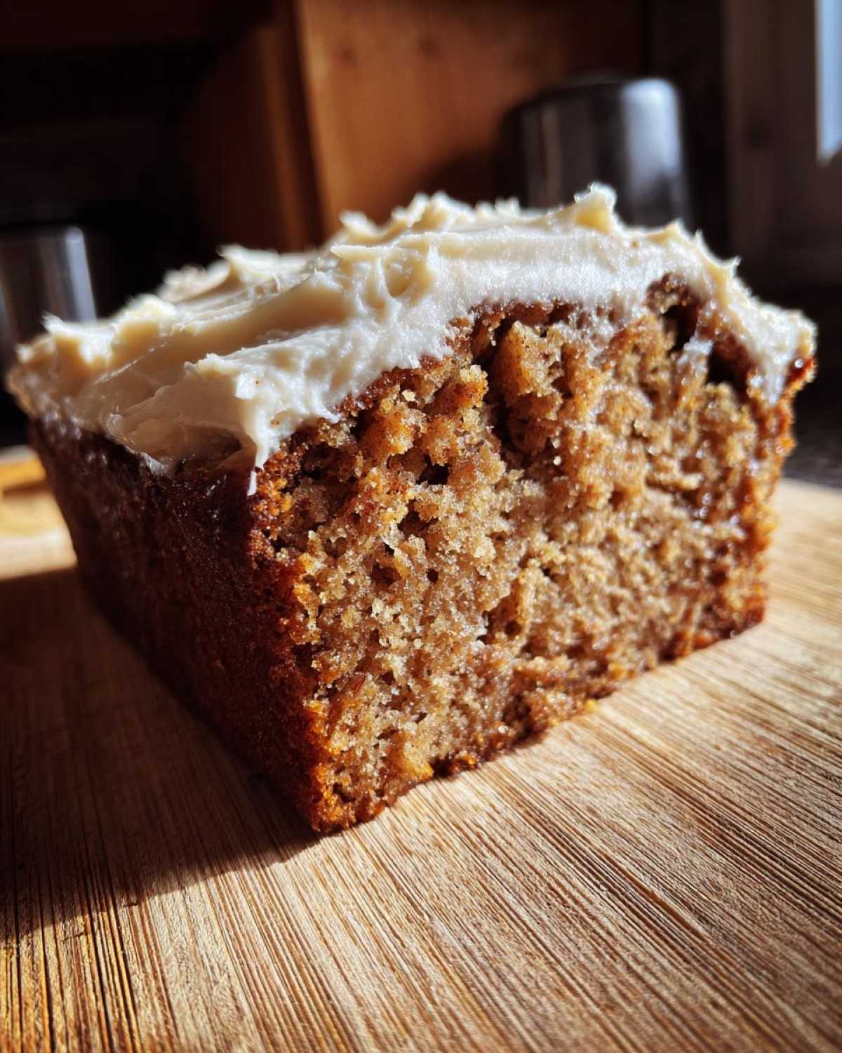 Close-up of a moist slice of applesauce cake topped with thick, white frosting, sitting on a wooden board.