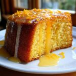 A close-up of a moist slice of orange juice cake drizzled with a thick, shiny glaze and topped with orange zest.