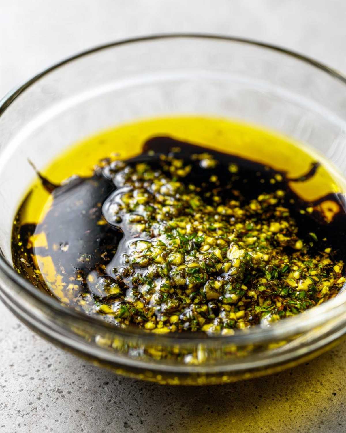 Close-up of olive oil bread dip ingredients: olive oil, balsamic vinegar, and minced herbs/garlic in a clear glass bowl.