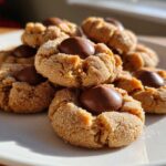 A tempting pile of freshly baked peanut butter blossom cookies, rolled in sugar and topped with chocolate kisses, sitting on a white plate.