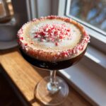 A festive peppermint espresso martini topped with foam and crushed candy canes, sitting by a sunny window.