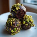 Close-up of rich, dark dubai chocolate balls coated in chopped green pistachios on a white plate.