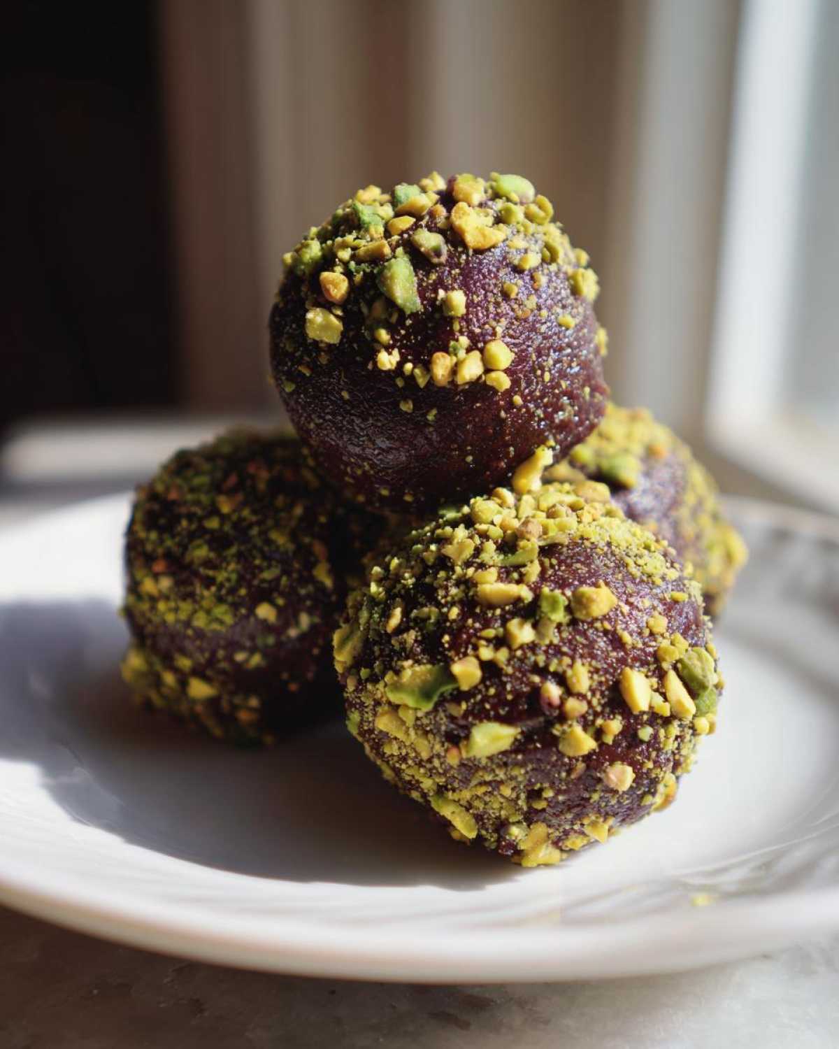 Close-up of rich, dark dubai chocolate balls rolled in bright green crushed pistachios, stacked on a white plate.