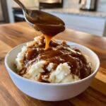 A spoonful of rich homemade brown gravy being poured over fluffy mashed potatoes in a white bowl.