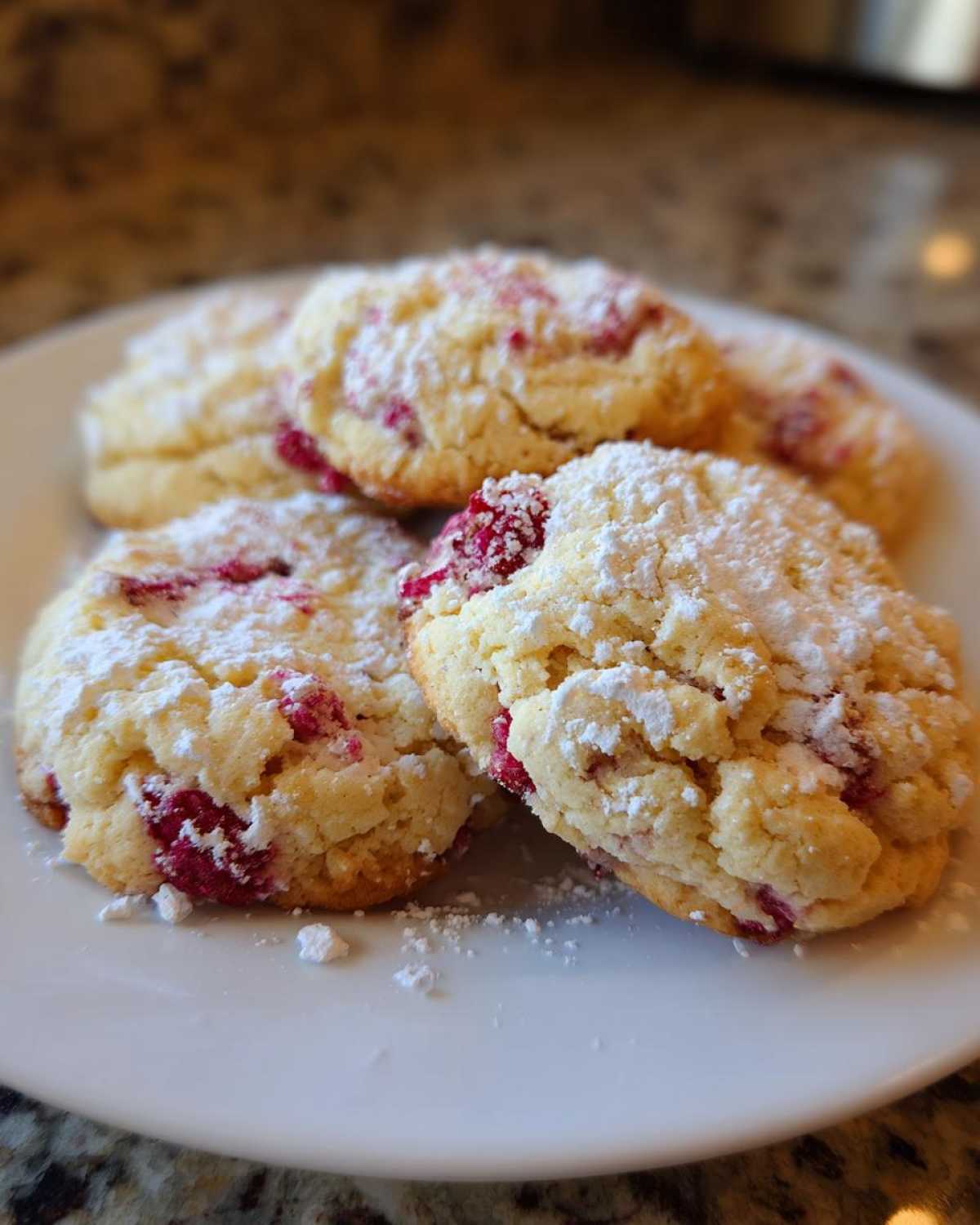 A close-up of several soft raspberry cheesecake cookies dusted heavily with powdered sugar on a white plate.