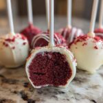A close-up view showing the inside texture of a red velvet cake pop dipped in white chocolate, sprinkled with red crumbs.