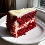 A bright slice of rich red velvet poke cake layered with cream cheese filling and topped with frosting.