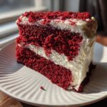 A thick slice of vibrant red velvet poke cake layered with white cream cheese frosting, sitting on a white plate.