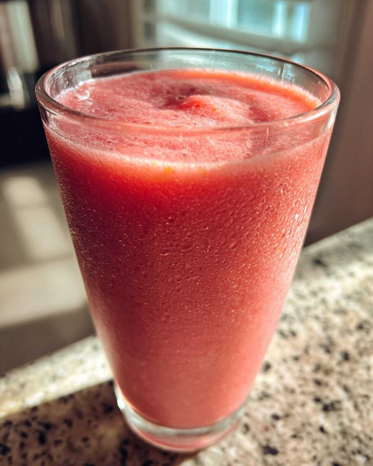 Close-up of a vibrant, frosty watermelon smoothie in a clear glass sitting on a granite countertop.