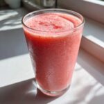 A tall glass filled with a bright pink, frosty watermelon smoothie, sitting on a white surface in bright sunlight.