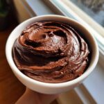 Close-up of thick, dark brown, swirled chocolate dip served in a small white bowl, held near a window.