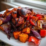 Close-up of roasted harissa chicken mixed with caramelized sweet potatoes, red bell peppers, and purple onions on a white plate.