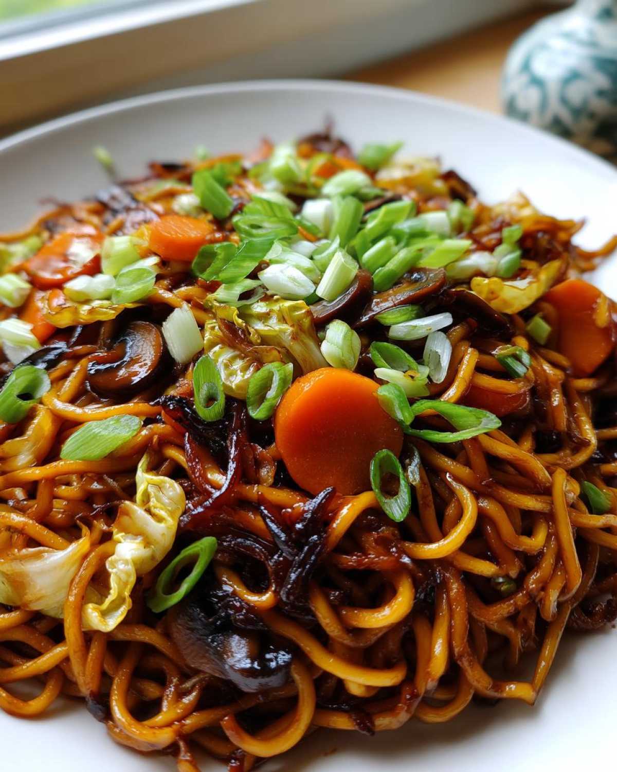A close-up of savory fried noodles coated in dark sauce, mixed with carrots, mushrooms, cabbage, and topped with green onions.
