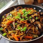 A close-up of savory fried noodles mixed with sautéed mushrooms and carrots, topped with fresh green onions, served in a black skillet.