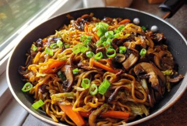 A close-up of savory fried noodles mixed with sautéed mushrooms and carrots, topped with fresh green onions, served in a black skillet.