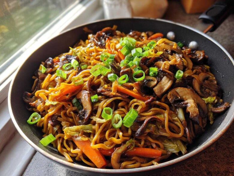 A close-up of savory fried noodles mixed with sautéed mushrooms and carrots, topped with fresh green onions, served in a black skillet.