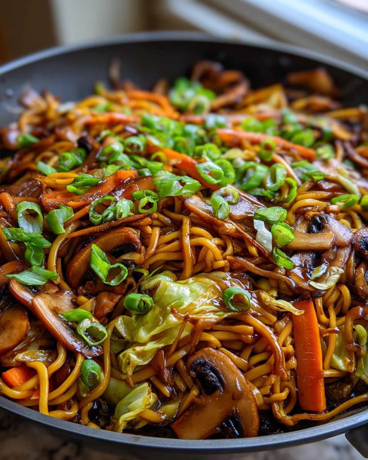 Close-up of savory fried noodles tossed with mushrooms, carrots, cabbage, and topped with green onions.