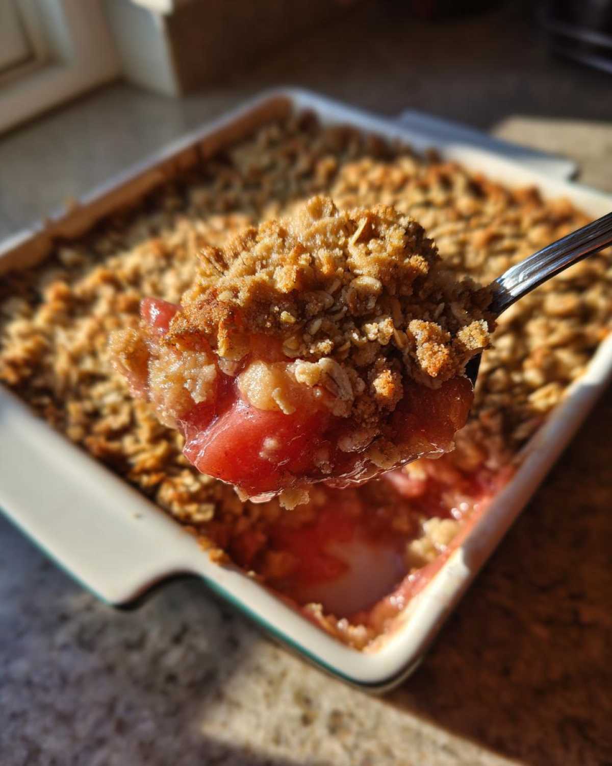 A spoonful of warm rhubarb crisp, showing the gooey fruit filling and crunchy oat topping, lifted from a baking dish.