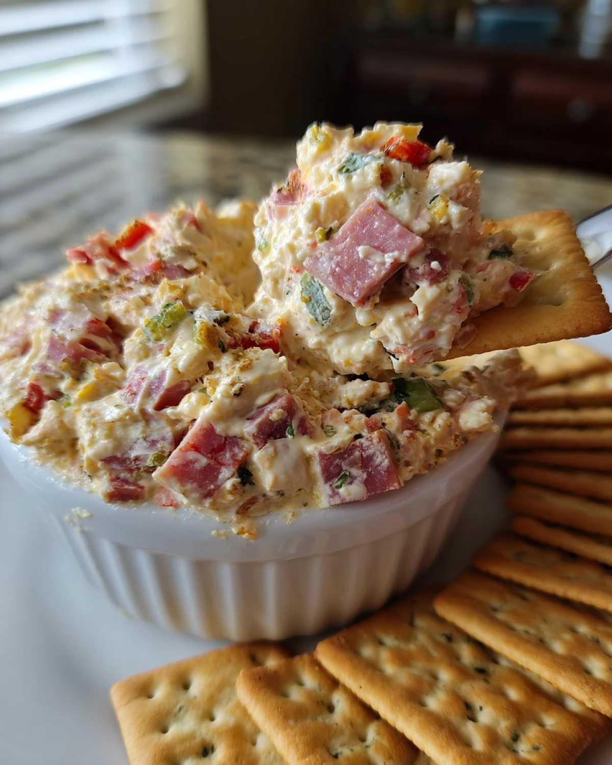 A spoonful of creamy hoagie dip filled with ham and peppers being scooped onto a square cracker.