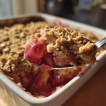 A spoonful of warm rhubarb crisp, showing the bubbly fruit filling and crunchy oat topping, lifted from a white baking dish.