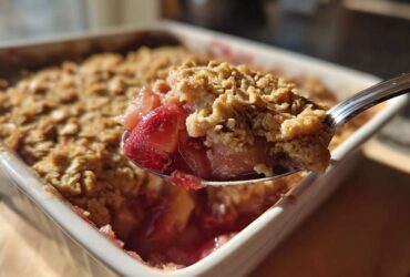 A spoonful of warm rhubarb crisp, showing the bubbly fruit filling and crunchy oat topping, lifted from a white baking dish.
