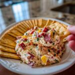 A hand scoops creamy hoagie dip loaded with meat and peppers onto a cracker.