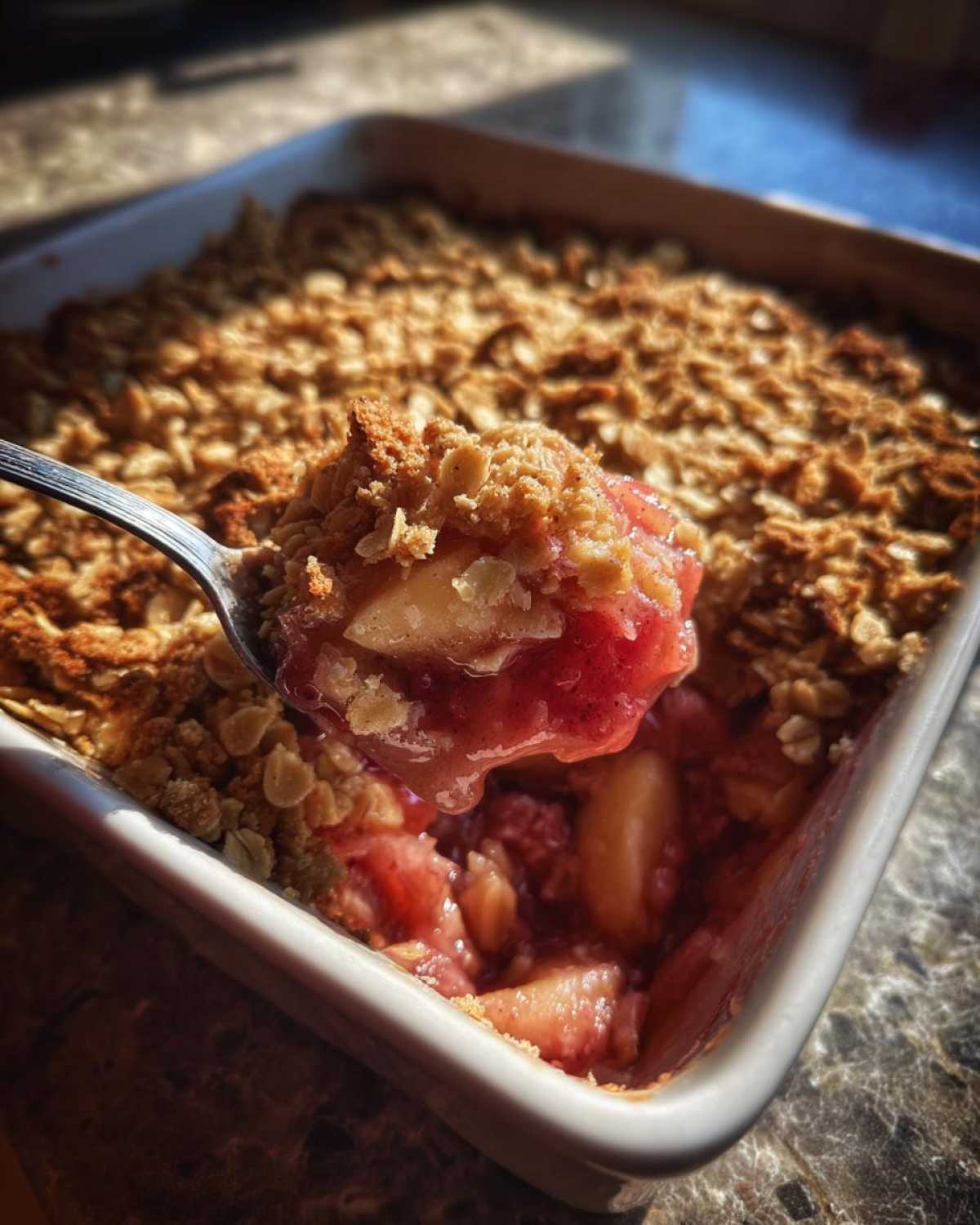A spoonful of warm, gooey rhubarb crisp being lifted from a white baking dish.