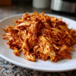 Close-up of flavorful, shredded chicken tinga coated in a rich, red adobo sauce served on a white plate.