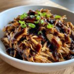 Close-up of tender, shredded crockpot teriyaki chicken coated in dark sauce and topped with green onions.