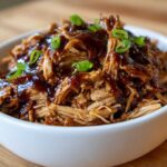 Close-up of shredded crockpot teriyaki chicken coated in thick sauce and topped with sliced green onions in a white bowl.