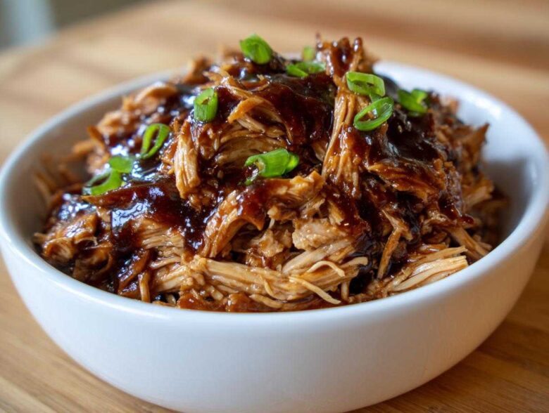 Close-up of shredded crockpot teriyaki chicken coated in thick sauce and topped with sliced green onions in a white bowl.