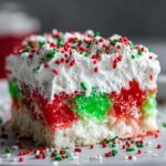 A close-up of a moist slice of christmas poke cake featuring layers of red and green gelatin, topped with whipped cream and holiday sprinkles.