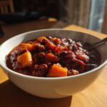 A steaming white bowl filled with rich, dark black bean chili featuring chunks of orange squash, sitting on a wooden table.