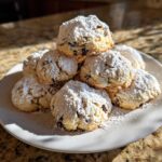 A mound of soft cannoli cookies, studded with chocolate chips and heavily dusted with powdered sugar, sitting on a white plate.