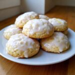 A close-up of soft ricotta cookies piled on a white plate, topped with a thick, sweet white glaze.