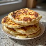 A stack of four golden, slightly charred homemade pita bread rounds resting on a white plate.