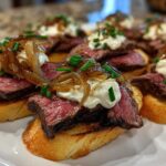 Close-up of several steak crostini appetizers topped with medium-rare steak, creamy sauce, caramelized onions, and chives.