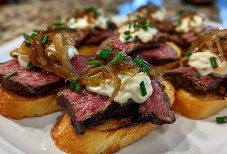 Close-up of several steak crostini appetizers topped with medium-rare steak, creamy sauce, caramelized onions, and chives.