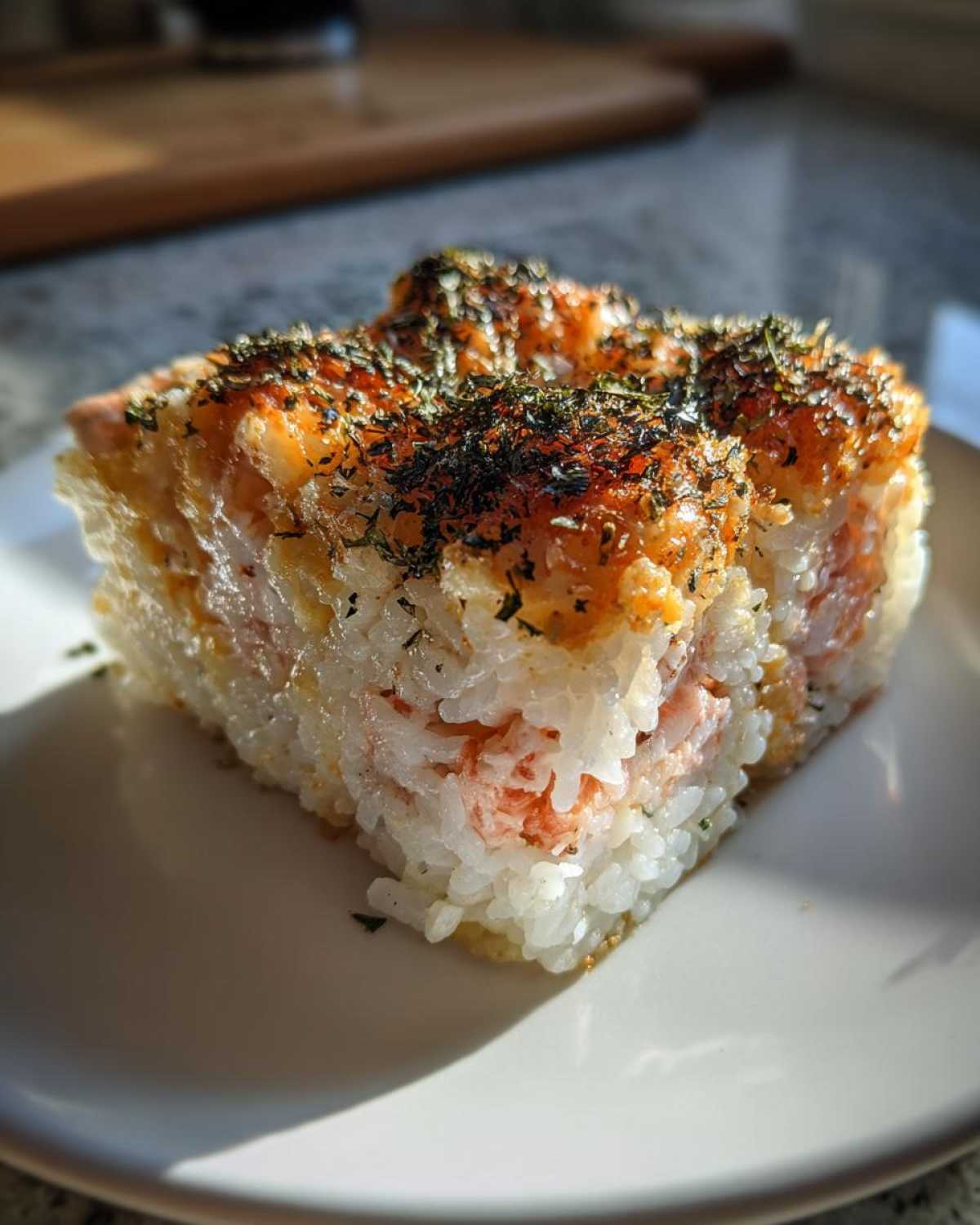 A square serving of baked sushi with creamy topping and seaweed flakes, showing layers of rice and filling.