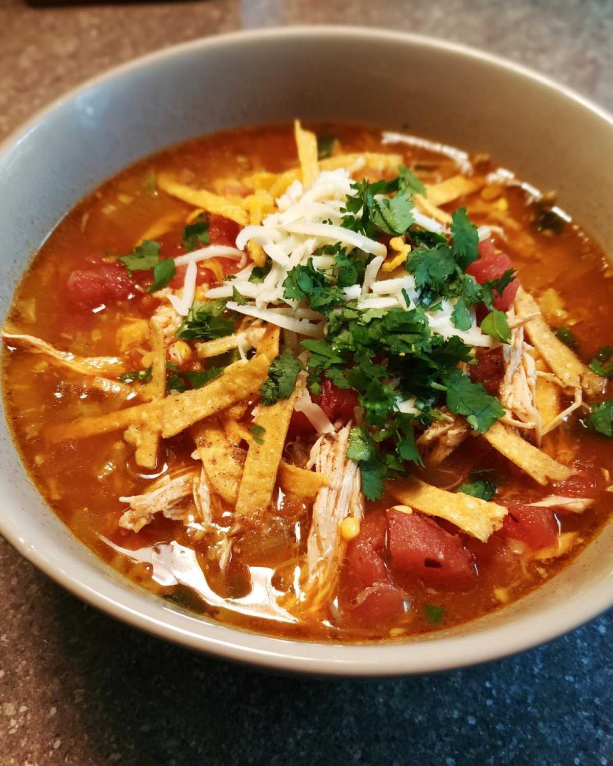 Close-up of a steaming bowl of tortilla soup recipe, topped with shredded chicken, cheese, cilantro, and crispy tortilla strips.