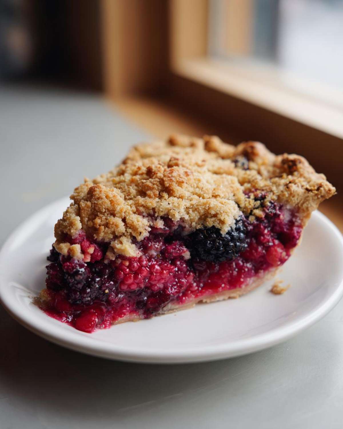 A close-up of a perfect slice of triple berry pie featuring a crumb topping served on a white plate.