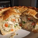 A close-up cross-section of a turkey pot pie recipe showing shredded turkey, peas, and carrots in a creamy sauce under a golden, flaky crust.