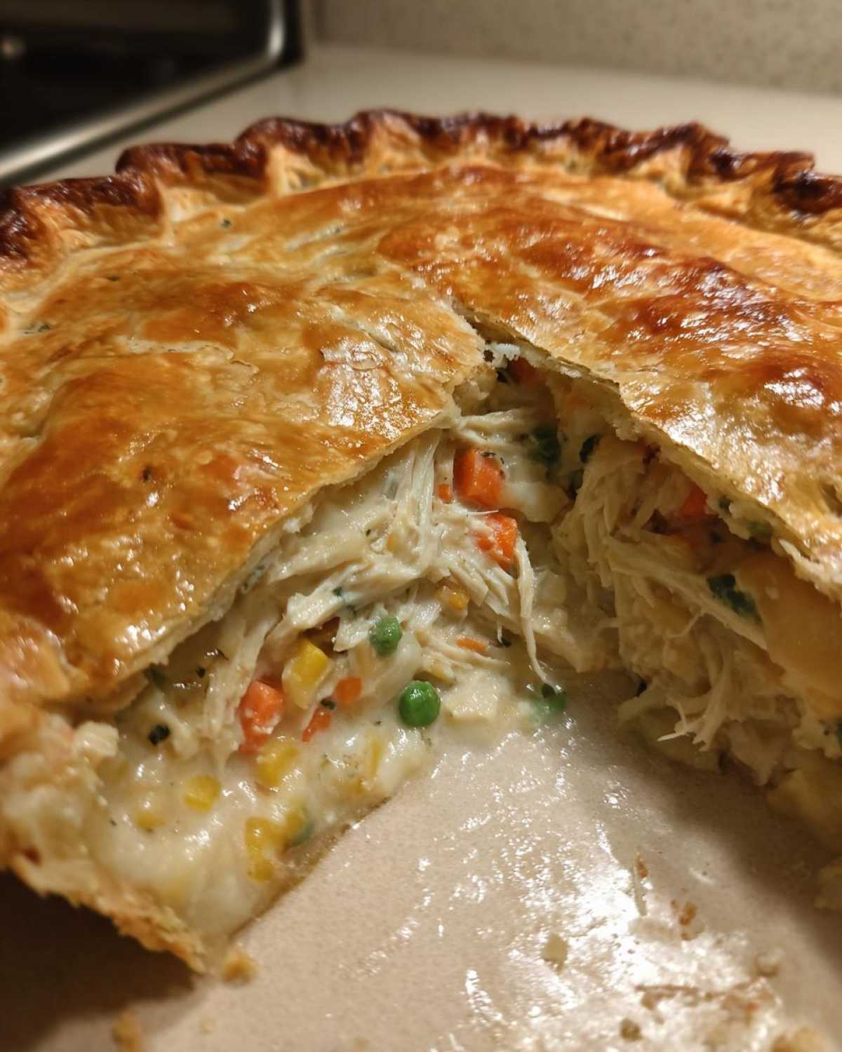 A close-up of a freshly baked turkey pot pie recipe with a slice cut out, revealing creamy filling, shredded turkey, carrots, and peas.