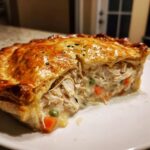 A close-up of a thick slice of turkey pot pie recipe showing shredded turkey, carrots, and peas in a creamy sauce under a golden, flaky crust.